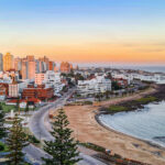 A imagem captura uma vista panorâmica da cidade costeira de Punta del Este, Uruguai, durante o pôr do sol. O horizonte urbano é adornado com edifícios altos e modernos, intercalados com estruturas mais baixas e tradicionais. A praia de areia se estende até a água azul clara, com algumas árvores e vegetação ao longo da costa. A atmosfera é calma e serena, com o céu pintado em tons de laranja e rosa.