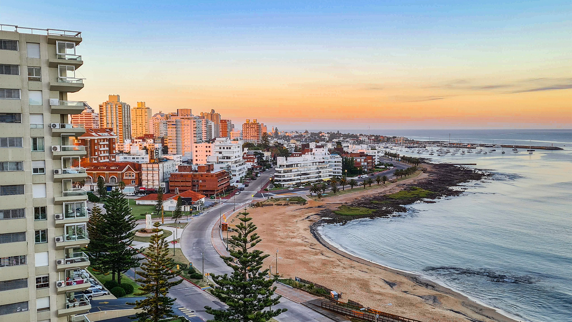 A imagem captura uma vista panorâmica da cidade costeira de Punta del Este, Uruguai, durante o pôr do sol. O horizonte urbano é adornado com edifícios altos e modernos, intercalados com estruturas mais baixas e tradicionais. A praia de areia se estende até a água azul clara, com algumas árvores e vegetação ao longo da costa. A atmosfera é calma e serena, com o céu pintado em tons de laranja e rosa.