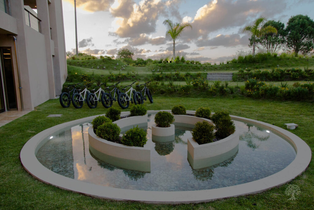 Alive health, noUruguai: Um jardim moderno com um pequeno lago circular no centro, cercado por plantas em vasos. O lago reflete o céu e a vegetação ao redor, criando uma atmosfera serena. Ao fundo, várias bicicletas estão estacionadas, sugerindo um ambiente de lazer e saúde. A cena é banhada por uma luz suave, indicando o pôr do sol, com palmeiras e gramados verdes exuberantes. A composição harmoniosa e a arquitetura limpa transmitem uma sensação de tranquilidade e bem-estar.