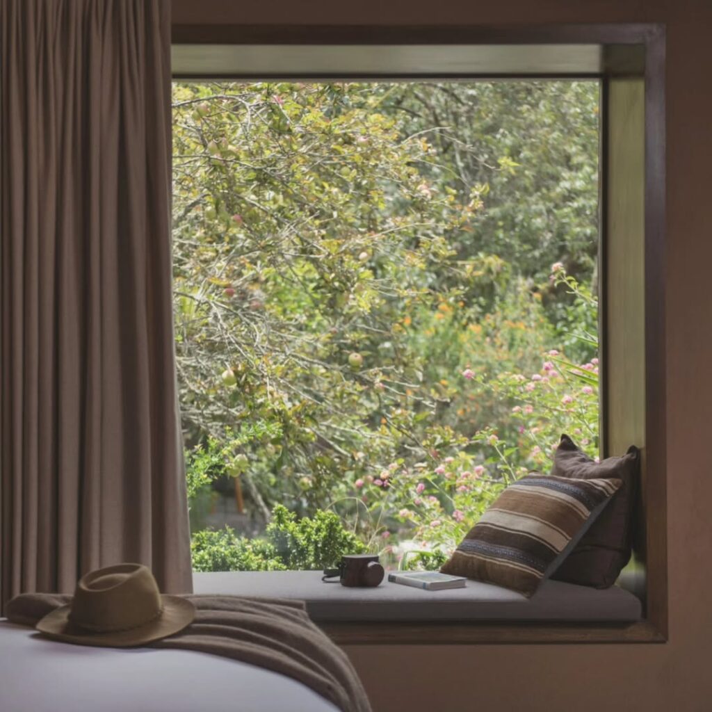 A imagem mostra uma janela em um quarto de hotel com vista para um jardim exuberante. A janela é emoldurada por cortinas bege e possui um banco com almofadas estriadas. No banco, há um chapéu de palha, um livro e uma xícara. O quarto tem um estilo moderno e acolhedor, com materiais como madeira e tecido.