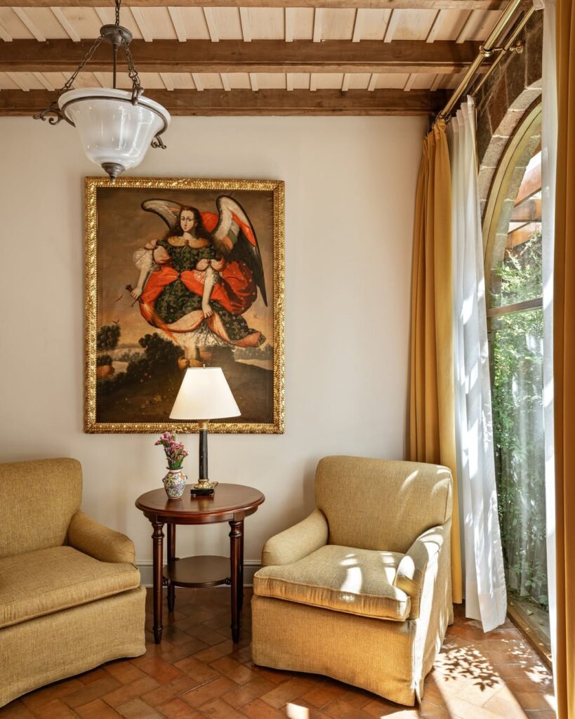A imagem retrata uma sala de estar no Belmond Hotel Monasterio, em Cusco, Peru. A sala possui móveis bege, incluindo duas poltronas confortáveis e uma mesa de centro de madeira. Uma pintura clássica de um anjo com asas é exibida proeminentemente na parede. A decoração inclui um abajur com uma lâmpada branca e um vaso com flores. A sala tem um chão de azulejos e uma janela arqueada com cortinas bege e brancas, permitindo a entrada de luz natural. Dica de hotel para um roteiro de viagem em Machu Picchu
