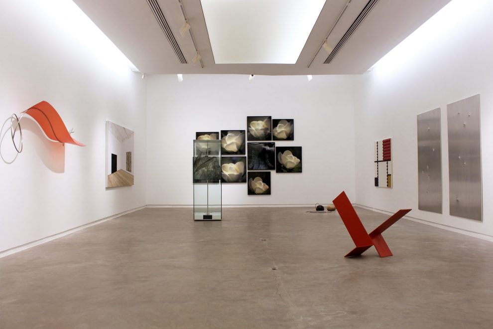 A imagem mostra uma exposição de arte moderna na galeria Silvia Cintra em 2020. O espaço é amplo e bem iluminado, com paredes brancas e piso de concreto. As peças de arte incluem esculturas abstratas e uma série de pinturas em preto e branco. As esculturas são feitas de materiais como metal e vidro, enquanto as pinturas são emolduradas e dispostas em uma grade.