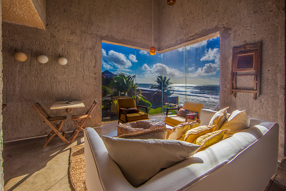 A imagem mostra uma sala de estar moderna com uma vista deslumbrante do mar no Kenoa Resort de São Miguel. A decoração é minimalista, utilizando materiais naturais e uma paleta de cores neutras. A sala possui um sofá branco com almofadas amarelas, uma mesa de centro de madeira, duas cadeiras de madeira e uma poltrona. A parede de vidro oferece uma vista panorâmica do oceano e da vegetação ao redor.