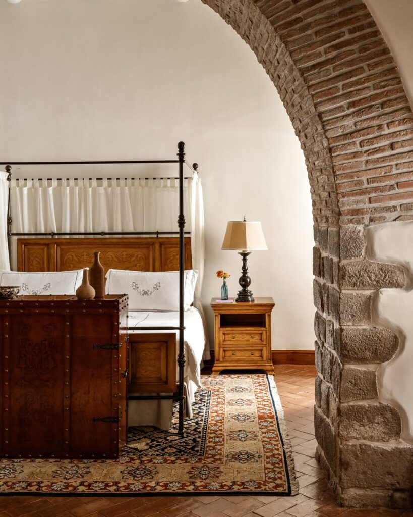 O quarto no Palacio Nazarenas em Machu Picchu, Peru, apresenta uma decoração clássica com um toque de elegância. A cama de dossel com cabeceira de madeira entalhada é o ponto focal, acompanhada por uma mesa de cabeceira de madeira com uma lâmpada. Um baú de madeira com ferragens e um tapete oriental adicionam calor ao espaço. A parede de tijolos e o arco de pedra conferem um charme rústico ao ambiente.