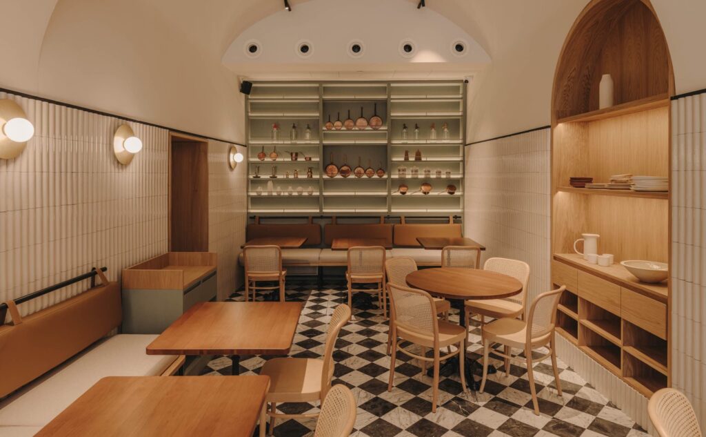 O restaurante Casa Heras no hotel em Girona Palau Fugit apresenta um design de interiores moderno e elegante. A paleta de cores predominante inclui tons de bege, marrom claro e branco, criando uma atmosfera quente e acolhedora. O espaço é mobiliado com mesas de madeira, cadeiras de vime e estantes de madeira que abrigam diversos utensílios e garrafas. O chão é adornado com um padrão xadrez preto e branco, adicionando um toque clássico ao ambiente. As paredes são revestidas com azulejos brancos e há iluminação suave proporcionada por luminárias embutidas e luminárias de parede. O design é caracterizado por uma combinação de materiais naturais como madeira e tecido, criando um espaço harmonioso e convidativo.
