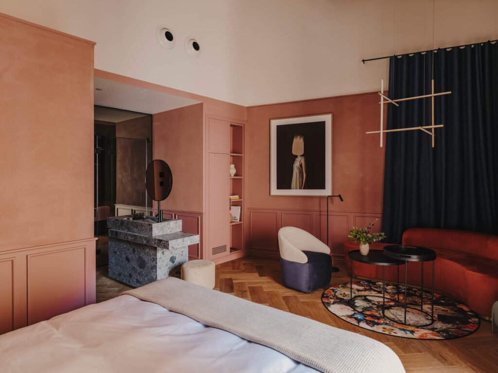 O quarto de hotel apresenta uma paleta de cores terrosas com paredes pintadas em um tom de rosa terroso. O mobiliário inclui uma cama com lençóis brancos, uma poltrona vermelha, uma mesa de centro preta e um pufe azul. Há um tapete circular com padrões florais e uma mesa de cabeceira com uma planta. A decoração é moderna e elegante, com um quadro artístico pendurado na parede e um espelho circular. O chão é de madeira, e há uma cortina azul escura cobrindo uma área de vidro. Hotel em Girona Palau Fugit