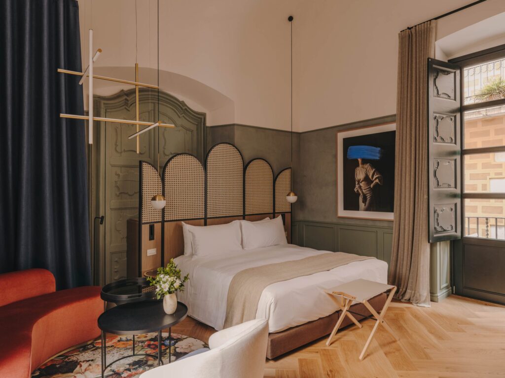 Hotel em Girona Palau Fugit. O quarto de hotel Palau Fugit na Espanha apresenta uma combinação de cores e estilos modernos. A cama é adornada com lençóis brancos impecáveis e tem uma cabeceira de madeira com detalhes em vime. Ao lado da cama, há uma poltrona vermelha e uma mesa de centro preta com um vaso de flores. A parede atrás da cama é decorada com um quadro de arte moderno. O quarto também possui um armário antigo e uma janela com cortinas bege que dão vista para um pátio externo.
