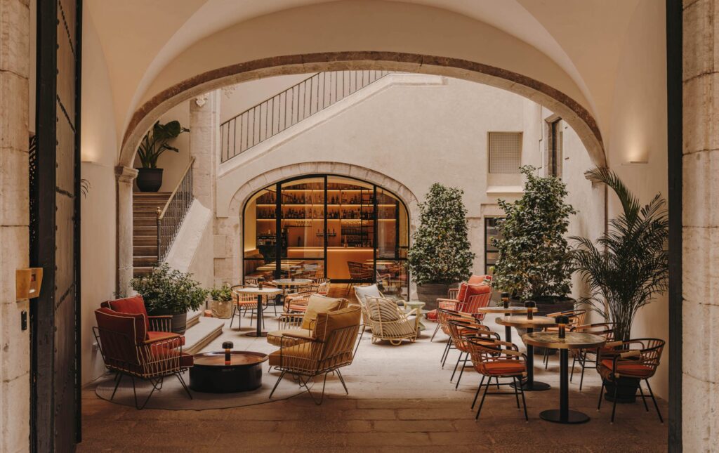 La Placeta, do Palau Fugit Hotel de Girona, na Espanha. A imagem mostra uma sala de estar moderna com detalhes históricos, combinando elementos contemporâneos com a herança do edifício. A sala possui móveis confortáveis, plantas em vasos e uma paleta de cores quentes. Há uma mistura de cadeiras e sofás com almofadas em tons de vermelho, laranja e bege. O ambiente é iluminado por luzes suaves e conta com um piso de madeira e paredes de pedra. A decoração inclui plantas verdes e um pátio interno com uma escada e uma área de refeições ao fundo.