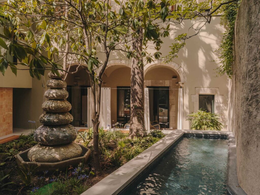 A imagem retrata um pátio interno sereno do hotel Palau Fugit, caracterizado por uma fonte de pedra e uma pequena piscina. O pátio é cercado por árvores e plantas exuberantes, criando uma atmosfera tranquila e natural. As paredes são pintadas em tons claros, complementando os elementos naturais. Arcos e colunas de pedra adicionam um toque de elegância arquitetônica.