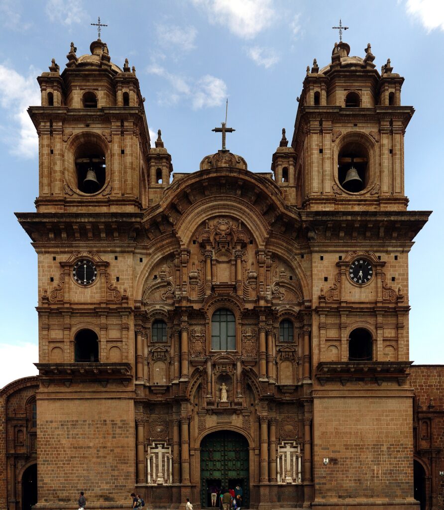 A imagem mostra a grandiosa fachada da Igreja da Companhia de Jesus em Cusco, Peru. A igreja apresenta um estilo barroco com duas torres imponentes, cada uma com um sino. A entrada principal é adornada com entalhes intricados e uma estátua de uma figura religiosa. O edifício é construído com pedra de cor marrom clara e possui janelas arqueadas e relógios em cada torre. Estilo barroco, area externa. 