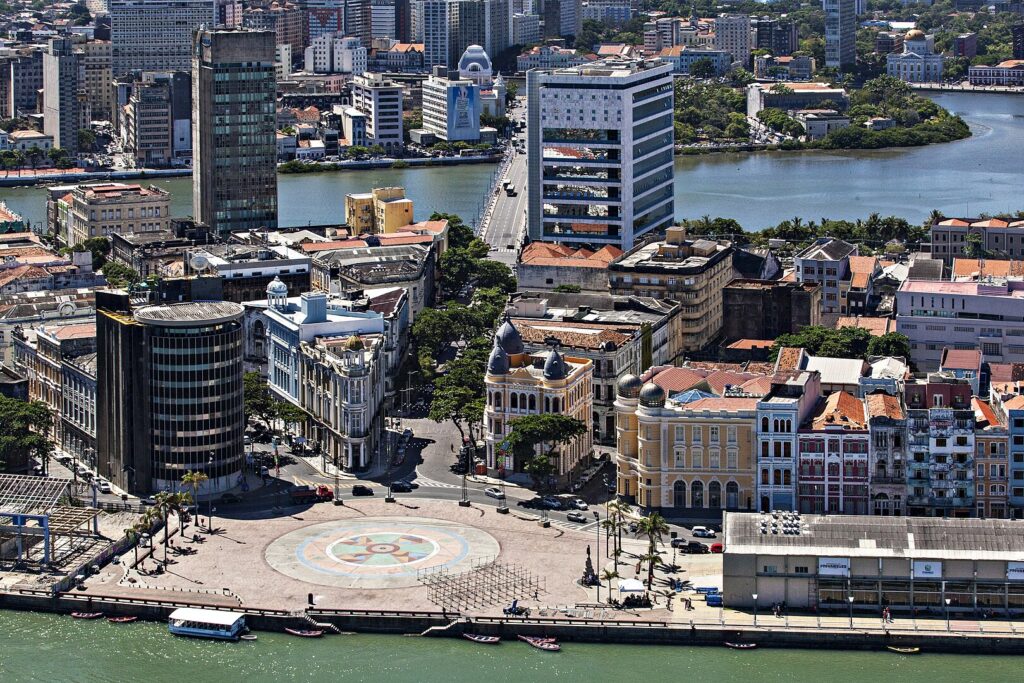 Praça urbana com edifícios históricos e modernos ao redor de um mosaico circular, a Rosa-dos-ventos, em tons de azul e verde. A cena é dominada por uma mistura de arquitetura colonial e contemporânea, com ruas movimentadas e um rio ao fundo. A atmosfera é vibrante e cultural, destacando a rica história e o design urbano de Recife.