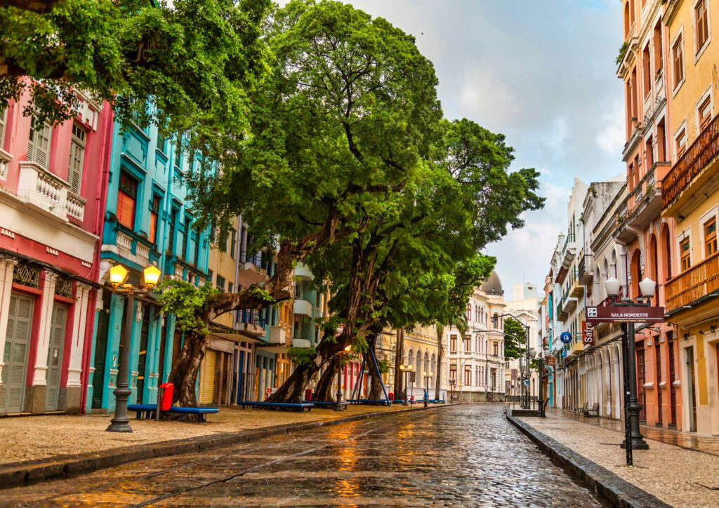 Rua pavimentada com pedra lioz molhada, refletindo a luz suave do entardecer. Edifícios coloridos em tons de azul, rosa e amarelo ladeiam a rua, com varandas e janelas ornamentadas. Grandes árvores verdes com troncos robustos e galhos espessos fornecem sombra e adicionam um toque de natureza à paisagem urbana. A atmosfera é tranquila e serena, com uma sensação de charme histórico e estética vibrante, característica do design colonial brasileiro. A composição equilibra a arquitetura e a vegetação, criando uma experiência visual harmoniosa e convidativa.