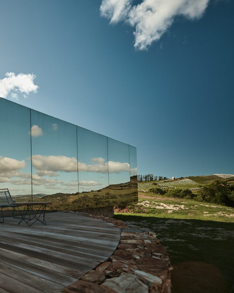 Hotel Sacromonte, no Uruguai. Uma varanda de madeira com uma mesa e cadeiras de metal sobre um deck de madeira, com vista para uma paisagem verdejante e colinas ondulantes. Ao fundo, um grande espelho refletivo captura o céu azul claro e nuvens brancas fofas, criando uma mistura harmoniosa de natureza e design moderno. A atmosfera é serena e convidativa, com uma sensação de tranquilidade e conexão com a natureza. O espelho, que reflete o céu e as nuvens, adiciona um toque de sofisticação e inovação arquitetônica.