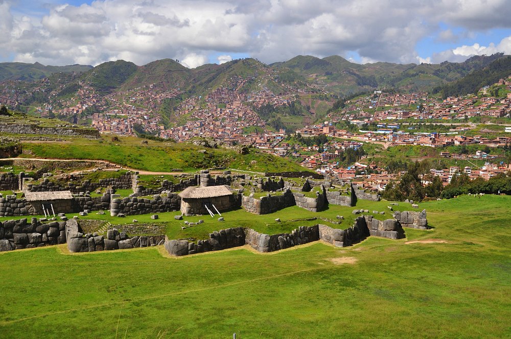 A imagem mostra as ruínas de Sacsayhuaman, um sítio arqueológico histórico, com uma vista panorâmica de uma cidade peruana ao fundo. As ruínas são compostas por estruturas de pedra meticulosamente construídas, cercadas por gramados verdes exuberantes. A paisagem urbana ao fundo apresenta uma mistura de edifícios e áreas verdes, com montanhas ao longe sob um céu nublado. machu picchu roteiro de viagem