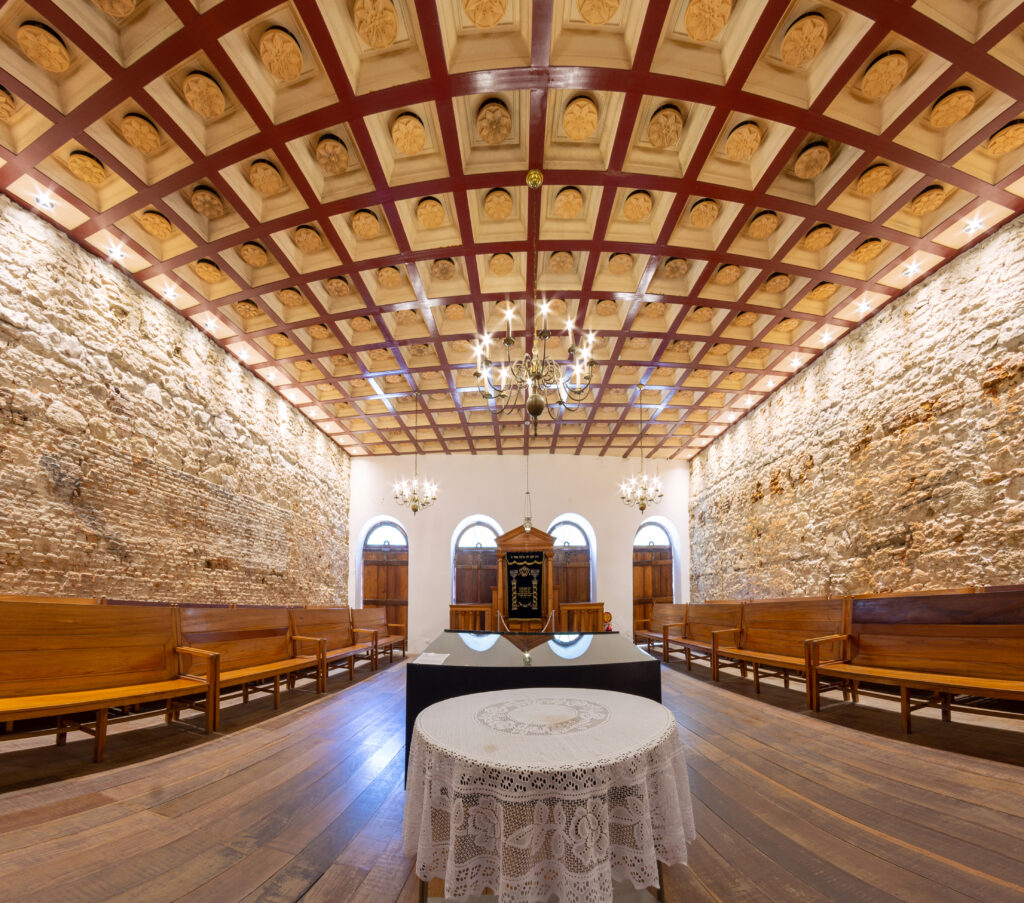 A imagem mostra o interior de uma sinagoga histórica, aKahal Zur Israel,com um teto ornamentado e paredes de pedra. O teto é adornado com padrões geométricos e decorações intricadas, destacando-se em tons marrons e brancos. A iluminação suave emanando de lustres adiciona uma atmosfera quente e acolhedora. No centro, há uma mesa coberta com uma toalha de renda branca, cercada por bancos de madeira. Ao fundo, pode-se ver uma arca da Torá, indicando a importância religiosa do espaço. A composição equilibrada e a atenção aos detalhes refletem um design cuidadoso, transmitindo uma sensação de reverência e tranquilidade. O que fazer emrecife