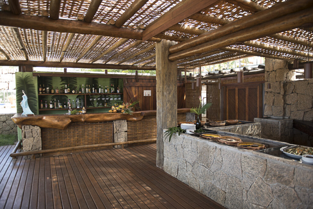 A imagem mostra um bar ao ar livre no Txai Resort, na Bahia, com uma decoração rústica que inclui um telhado de bambu, paredes de pedra e madeira, e uma variedade de garrafas de bebidas. O bar é adornado com plantas e flores, criando uma atmosfera acolhedora e natural. Diferenças entre Resort e Hotel