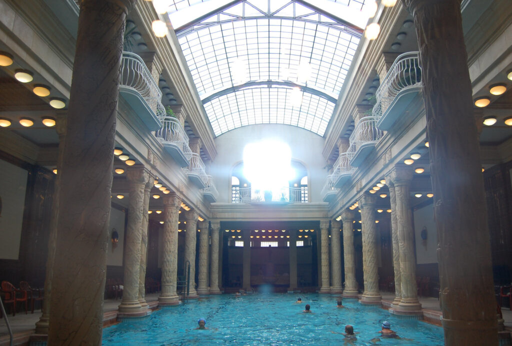 espaço tradicional de banho turco. A imagem mostra uma piscina interna suntuosa com colunas de mármore e uma claraboia que permite a entrada de luz natural. O ambiente é adornado com balustradas e iluminação circular, criando uma atmosfera elegante e serena. A piscina é cercada por colunas altas e há pessoas nadando na água.