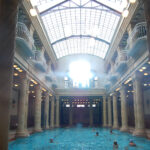 espaço tradicional de banho turco. A imagem mostra uma piscina interna suntuosa com colunas de mármore e uma claraboia que permite a entrada de luz natural. O ambiente é adornado com balustradas e iluminação circular, criando uma atmosfera elegante e serena. A piscina é cercada por colunas altas e há pessoas nadando na água.
