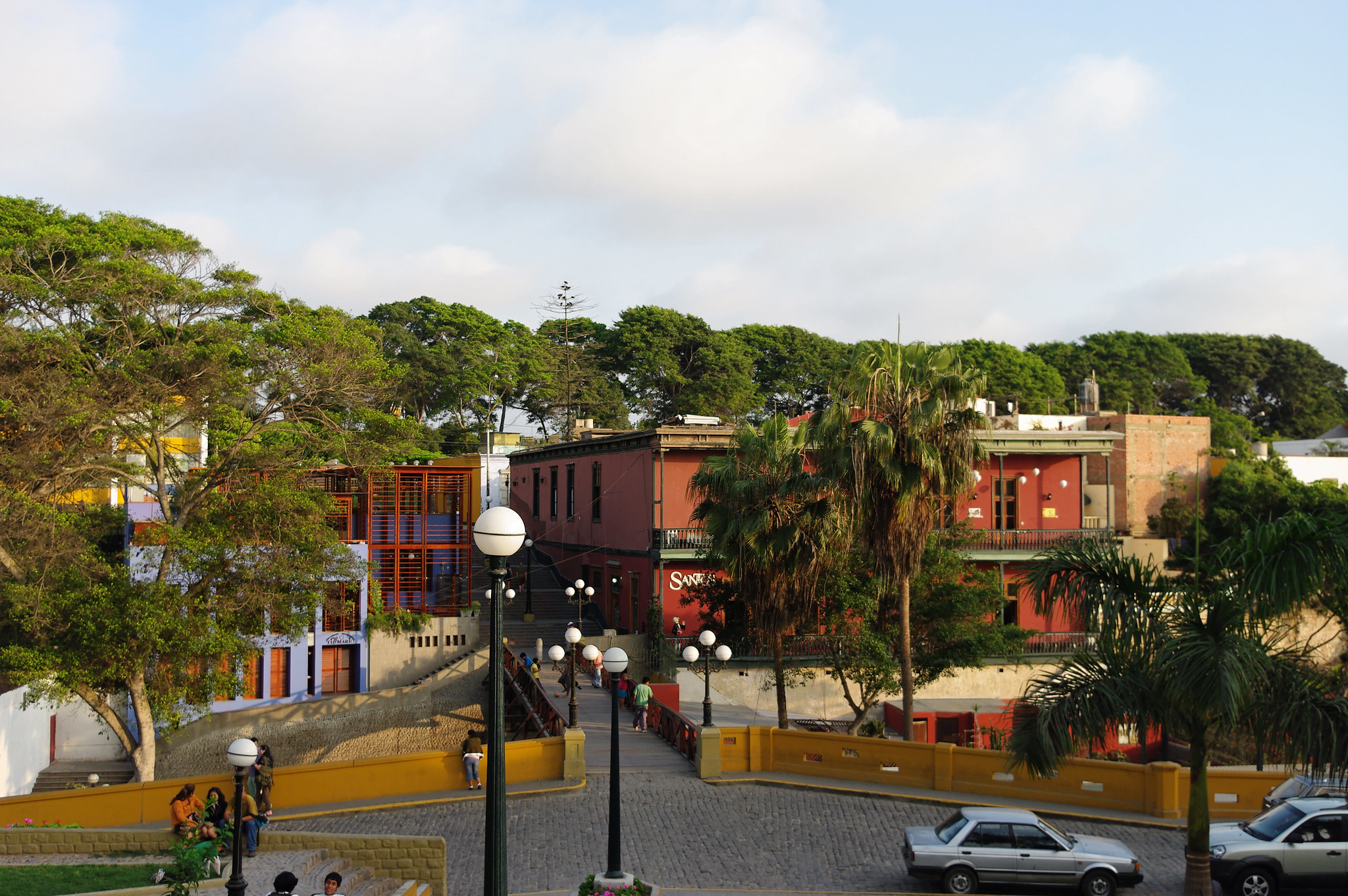 A imagem retrata uma rua de paralelepípedos no bairro de Barranco, conhecido como o distrito das artes em Lima, Peru. Os edifícios são pintados em tons vibrantes de vermelho e laranja, contrastando com a vegetação exuberante das árvores e palmeiras. A área parece ser uma mistura de espaços comerciais e residenciais, com um restaurante visível e pessoas caminhando pela rua. A decoração é moderna e colorida, com elementos únicos como a iluminação de rua e a arte nas