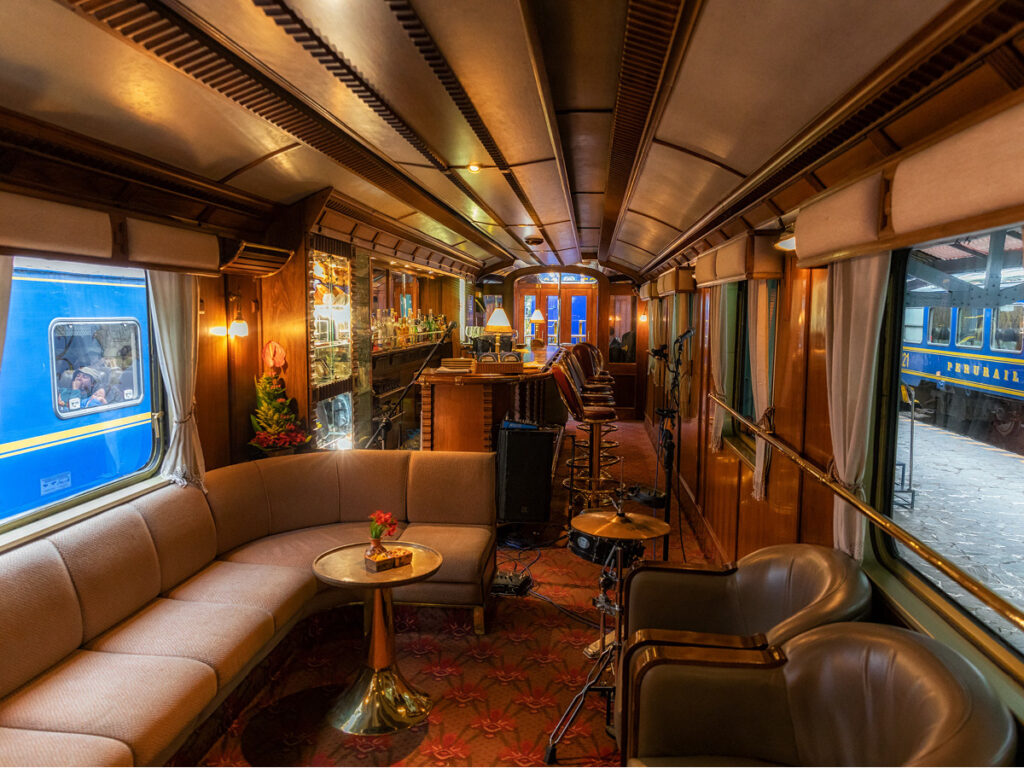 A imagem mostra o interior de um vagão de trem luxuoso, com uma paleta de cores quentes e acolhedoras. O ambiente é decorado com móveis de madeira, sofás confortáveis e um bar bem equipado. A decoração inclui elementos clássicos, como cortinas pesadas, tapetes de padrão elaborado e iluminação suave. O vagão de trem é parte do Belmond Hiram Bingham, que liga cidades do Peru a Machu Picchu.