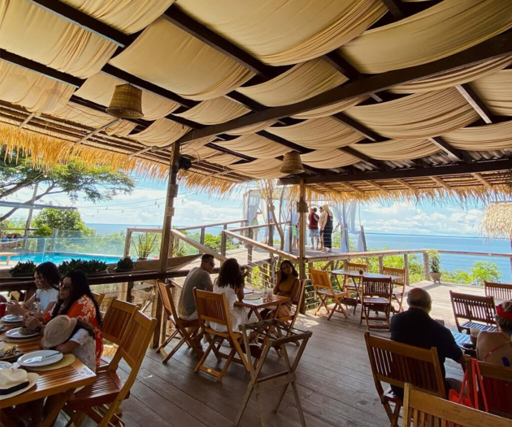 A imagem mostra a área externa da Casa do Saulo em Santarém, com um deck de madeira coberto por um telhado de palha. Várias mesas e cadeiras de madeira estão dispostas, onde pessoas estão sentadas e conversando. Ao fundo, há uma vista deslumbrante do oceano e uma piscina. O ambiente é relaxado e tropical, com decoração rústica e elementos naturais.