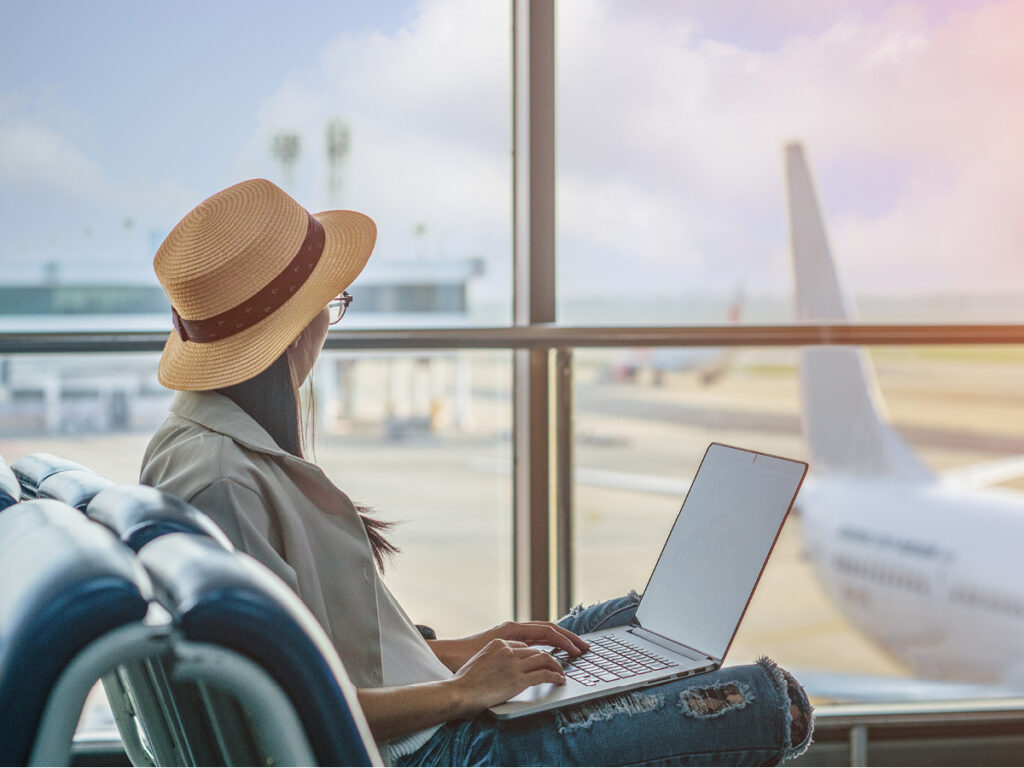 A imagem mostra uma mulher usando um chapéu de palha e óculos, sentada em uma cadeira de couro em um aeroporto. Ela está trabalhando em um laptop enquanto espera por seu voo. Ao fundo, há uma janela grande com vista para a pista do aeroporto, onde um avião está estacionado. A cena é iluminada por luz natural, criando uma atmosfera calma e produtiva.