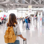 A imagem mostra uma mulher com uma mochila laranja em um terminal de aeroporto. Ela está de costas, olhando para um painel de informações. O terminal é espaçoso com um teto alto e iluminação brilhante. Há várias pessoas ao fundo, algumas sentadas em bancos e outras caminhando. A decoração é moderna com elementos industriais. Voos low cost