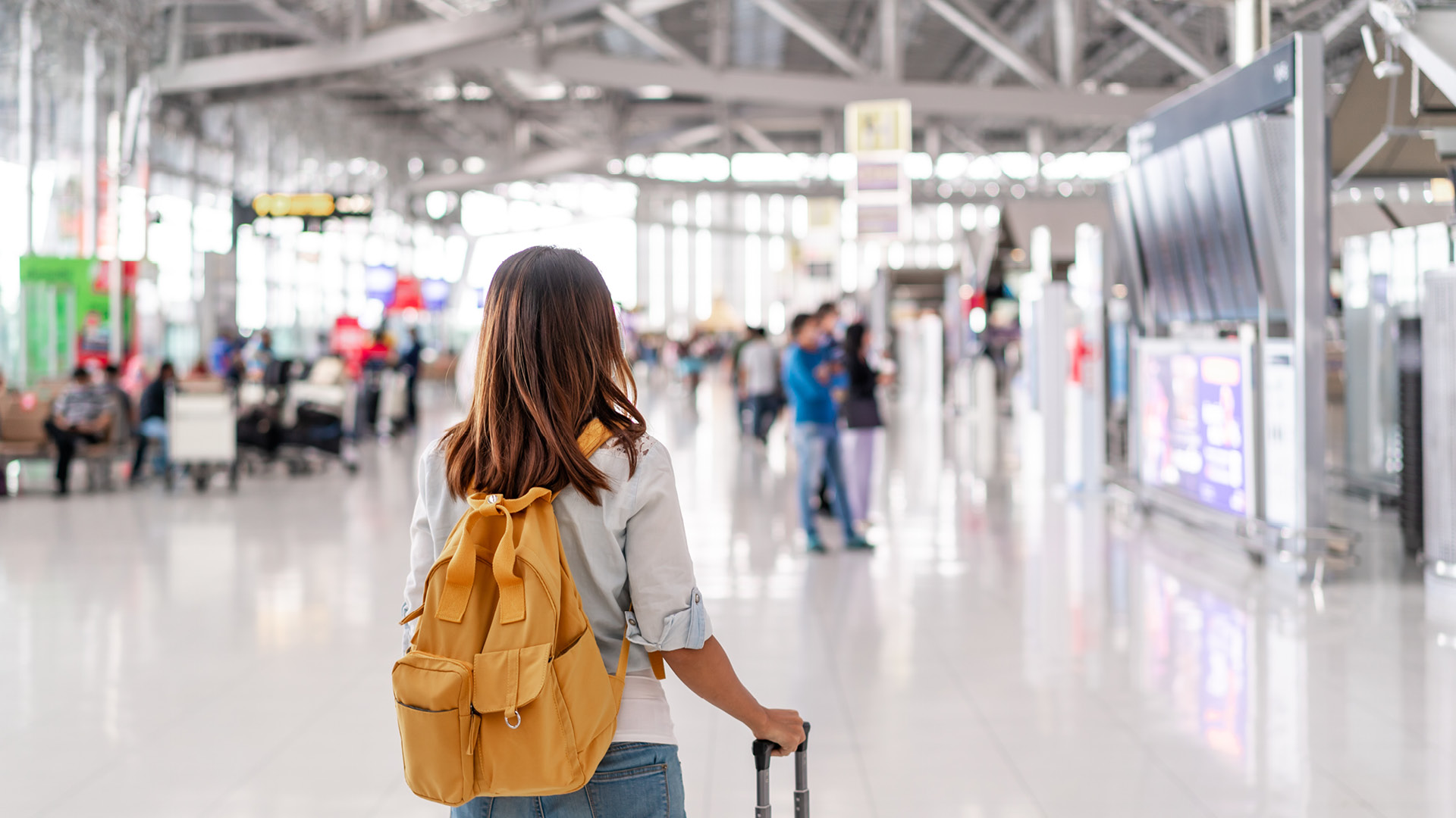 A imagem mostra uma mulher com uma mochila laranja em um terminal de aeroporto. Ela está de costas, olhando para um painel de informações. O terminal é espaçoso com um teto alto e iluminação brilhante. Há várias pessoas ao fundo, algumas sentadas em bancos e outras caminhando. A decoração é moderna com elementos industriais. Voos low cost