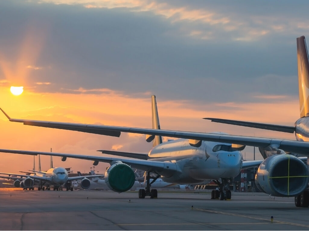 A imagem retrata um aeroporto durante o pôr do sol, com vários aviões estacionados na pista. O céu está pintado com tons de laranja e azul, criando um cenário sereno e tranquilo. Os aviões são modernos e bem conservados, com detalhes metálicos e cores que contrastam com o céu colorido. Vos low cost