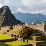A imagem mostra a impressionante paisagem de Machu Picchu, um sítio arqueológico histórico localizado nas montanhas dos Andes no Peru. As estruturas de pedra antigas e bem preservadas estão aninhadas em meio a uma vegetação exuberante, com montanhas imponentes ao fundo. O céu está parcialmente nublado, adicionando um clima dramático à cena. Machu Picchu roteiro de viagem