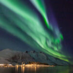 A imagem captura uma vista deslumbrante da aurora boreal em Tromsø, na Noruega. O céu noturno é iluminado por luzes verdes e roxas brilhantes da aurora boreal, refletindo nas águas calmas abaixo. Montanhas cobertas de neve e uma vila com luzes brilhantes ao longe acrescentam à beleza cênica.