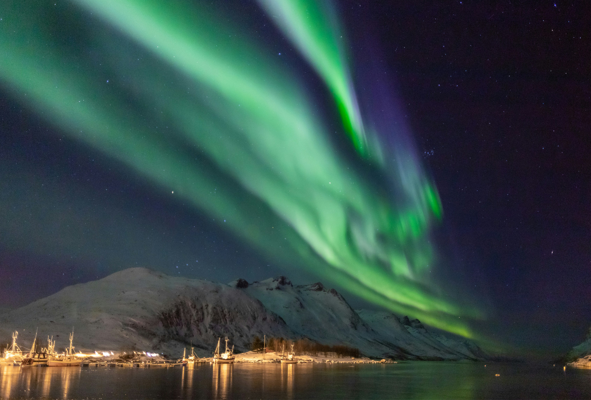A imagem captura uma vista deslumbrante da aurora boreal em Tromsø, na Noruega. O céu noturno é iluminado por luzes verdes e roxas brilhantes da aurora boreal, refletindo nas águas calmas abaixo. Montanhas cobertas de neve e uma vila com luzes brilhantes ao longe acrescentam à beleza cênica.