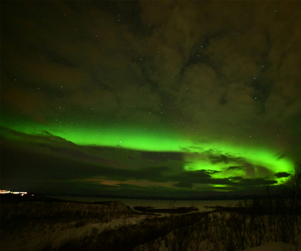 A imagem captura uma vista deslumbrante da aurora boreal em Abisko, Suécia. O céu noturno é iluminado por uma aurora verde brilhante, com nuvens escuras adicionando profundidade à cena. A paisagem abaixo está coberta de neve, proporcionando um contraste marcante com o espetáculo celestial acima.