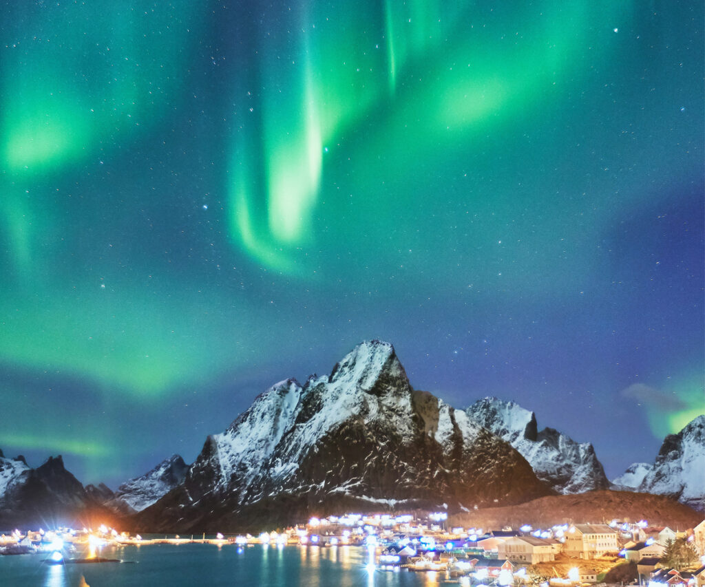 A imagem captura uma vista deslumbrante da aurora boreal iluminando o céu noturno sobre as ilhas de Loften na Noruega. As luzes verdes vibrantes da aurora contrastam lindamente com o céu escuro e as montanhas cobertas de neve ao fundo. À frente, uma pequena cidade costeira com luzes brilhantes adiciona um toque de calor à cena. A água calma reflete as luzes da cidade e a aurora, criando um reflexo sereno.