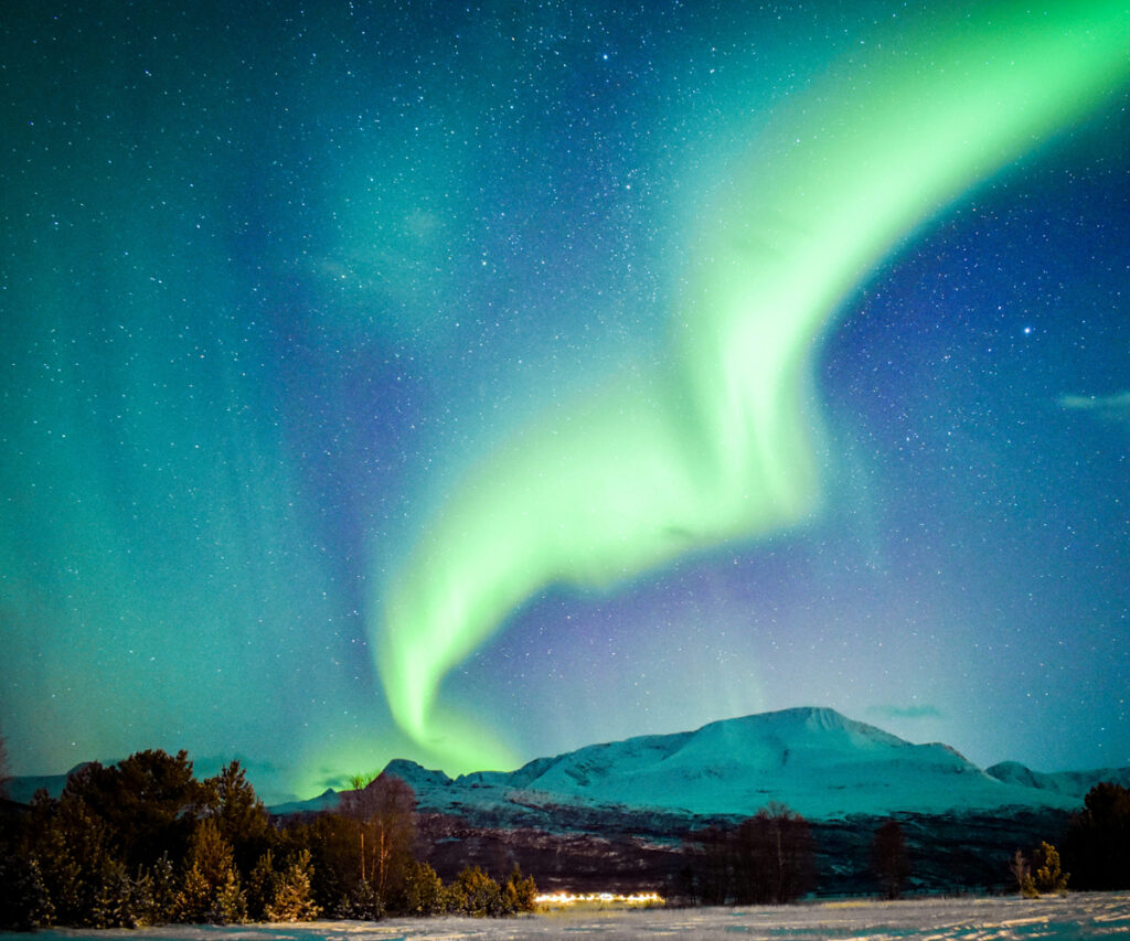 A imagem captura uma vista deslumbrante da Aurora Boreal em Tromsø, Noruega. O céu noturno é adornado com uma exibição vibrante de luzes verdes e azuis, criando um espetáculo natural sobre as montanhas cobertas de neve. A paisagem é serena, com árvores e uma pequena área habitada ao fundo.