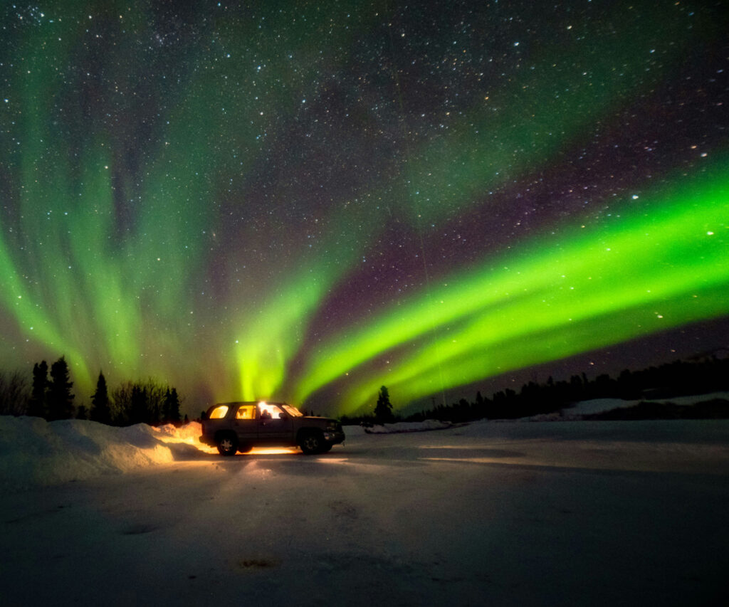 A imagem captura uma vista deslumbrante da aurora boreal em Fairbanks, Estados Unidos. O céu noturno é iluminado por vibrantes tons de verde e amarelo, criando um espetáculo natural sobre a paisagem nevada. Um carro está estacionado na estrada coberta de neve, com os faróis acesos, adicionando um toque de luz artificial à cena. Ao fundo, árvores e uma linha do horizonte distante são visíveis, realçando a beleza serena do ambiente. O Alasca é um dos principais destinos para ver aurora boreal na américa do norte