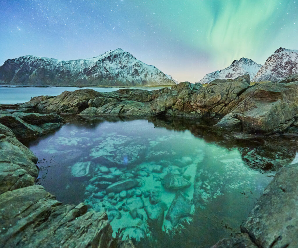 A imagem captura uma vista deslumbrante de um dos principais destinos para ver a aurora boreal, as Ilhas Lofoten na Noruega. O céu noturno é adornado com uma exibição vibrante de luzes verdes e azuis, refletidas em uma pequena piscina de água cercada por rochas. Ao fundo, montanhas cobertas de neve se erguem majestosamente, aumentando a beleza cênica do local.