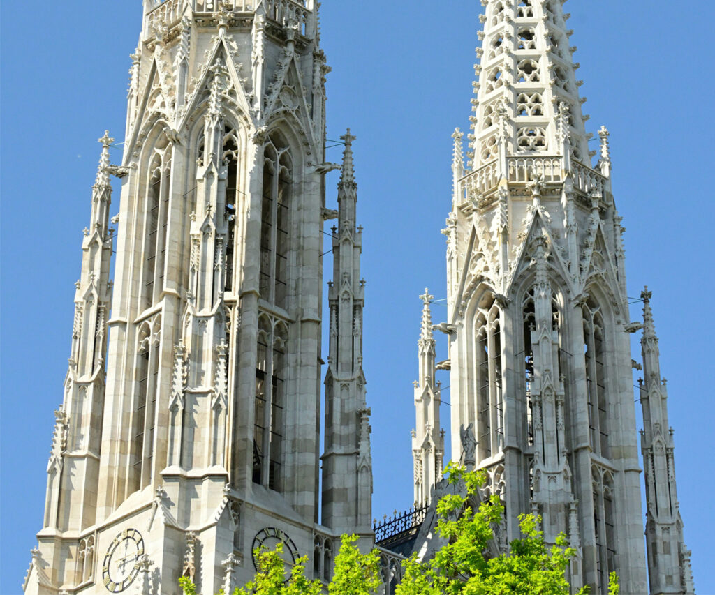A imagem apresenta as torres góticas da Catedral de Santo Estêvão I, destacando-se contra um céu azul claro. As torres são adornadas com detalhes intricados e janelas em arco, exibindo a arquitetura gótica clássica. A catedral é cercada por árvores verdes exuberantes, adicionando um toque de natureza à estrutura histórica.