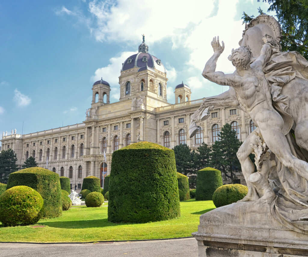 Dicas de viagem para Viena: A imagem mostra o Museu da História Nacional de Viena com sua arquitetura grandiosa e detalhada. No primeiro plano, há uma estátua de uma figura mitológica e um jardim bem cuidado com arbustos e árvores. O céu está azul com algumas nuvens, criando um ambiente sereno e histórico.