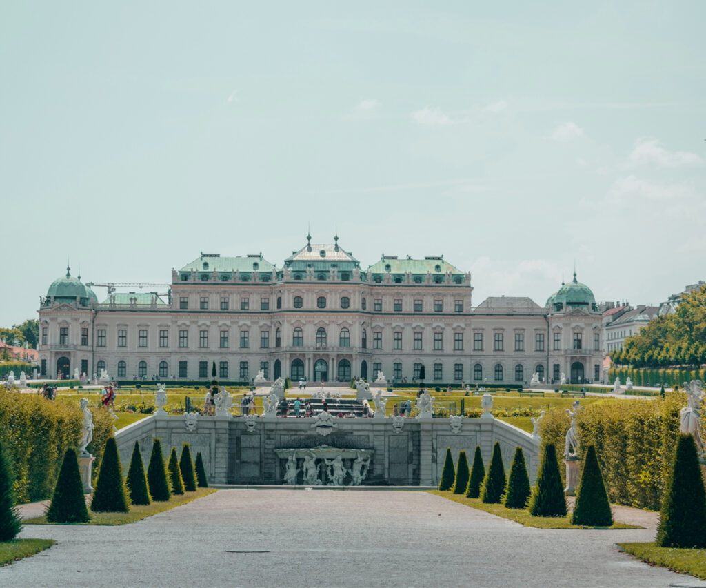 Dicas de viagem para Viena: A imagem retrata o majestoso Palácio Belvedere de Viena, um marco histórico com uma arquitetura barroca impressionante. O palácio é cercado por jardins bem cuidados, com estátuas ornamentadas e árvores bem podadas. A entrada principal é marcada por um caminho largo que leva até o edifício principal, destacando a grandiosidade e a elegância do local.