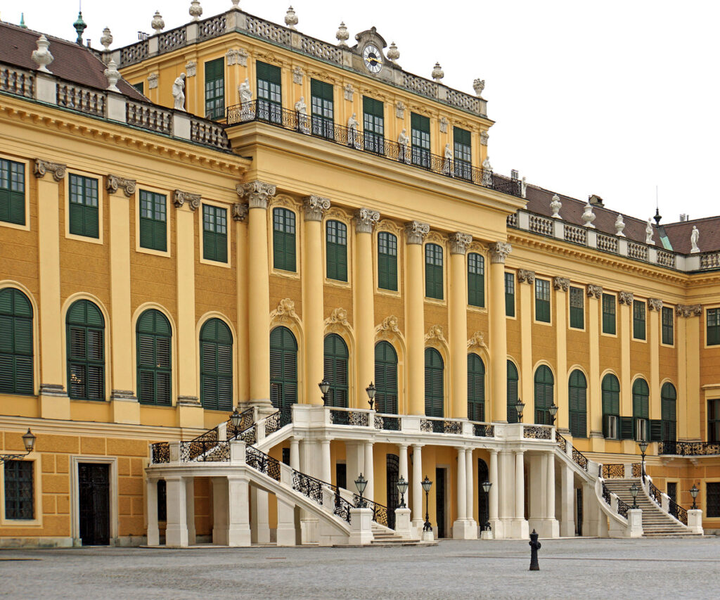 A imagem mostra a grandiosa fachada do Palácio Schönbrunn em Viena, Áustria. O edifício é pintado em um tom amarelo vibrante com janelas verdes e detalhes ornamentados. A arquitetura é caracterizada por colunas brancas e escadas que levam às entradas principais. O pátio em frente ao palácio é pavimentado com paralelepípedos, e há um poste de luz próximo à entrada.