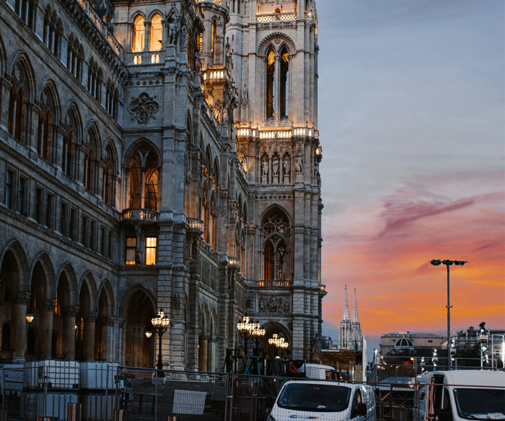 Dicas de viagem para Viena A imagem captura a Prefeitura de Viena, um edifício gótico renascentista, iluminado ao pôr do sol. A fachada do edifício é adornada com janelas arqueadas e detalhes ornamentados. Na frente do edifício, há vários veículos estacionados, incluindo vans e carros. O céu ao fundo está pintado em tons de laranja e rosa, criando um contraste dramático com a arquitetura histórica.