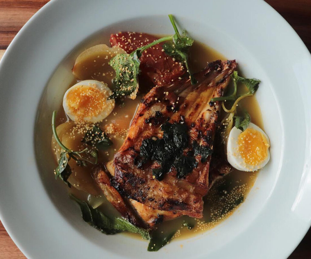 A imagem apresenta uma deliciosa caldeirada de peixe servida no restaurante Caxiri da gastronomia da amazônia, com decoração moderna. O prato é apresentado em uma tigela branca, com pedaços de peixe grelhado, ovos cozidos, vegetais e temperos. A mesa de madeira adiciona um toque rústico ao ambiente elegante.