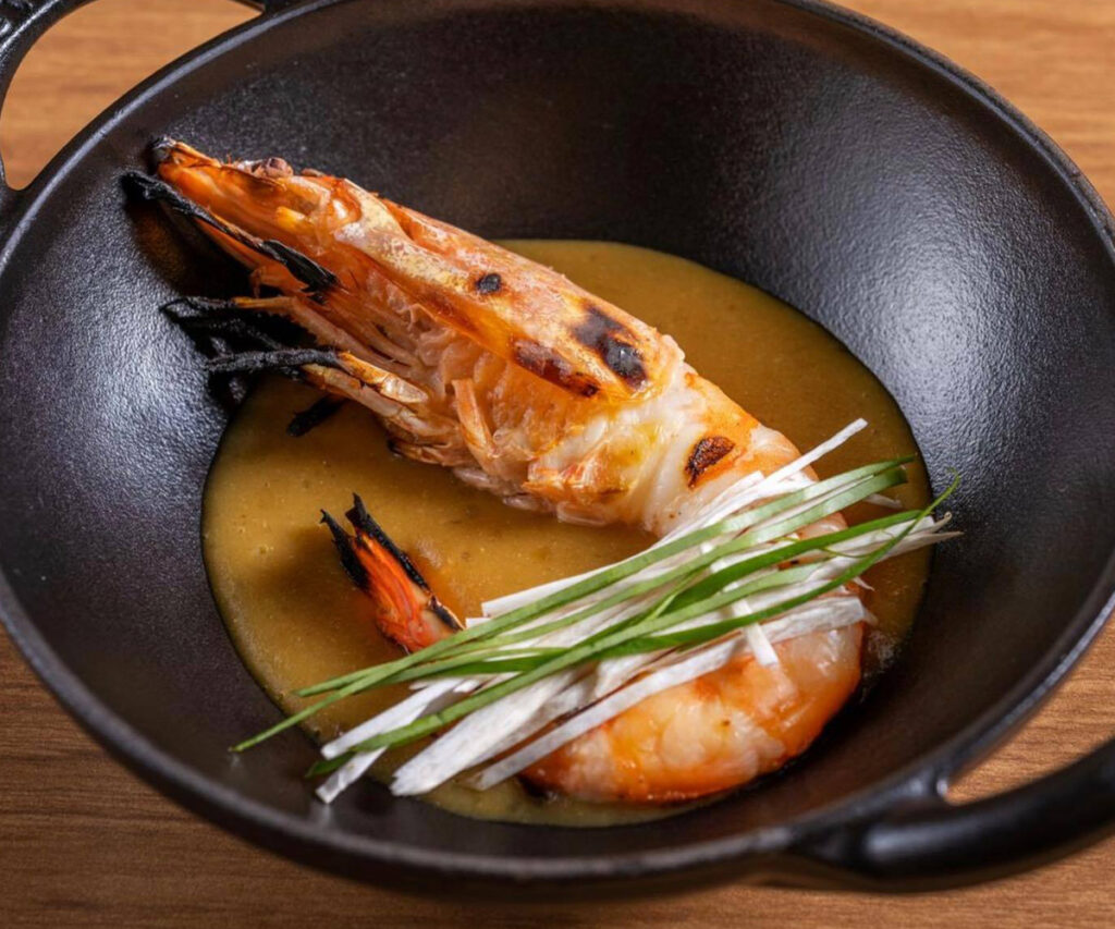 A imagem apresenta um prato gourmet do Restaurante Banzeiro, caracterizado por um camarão assado com molho cremoso em uma tigela preta. O prato é decorado com cebolinha verde e fatias de vegetais, servido em uma mesa de madeira.