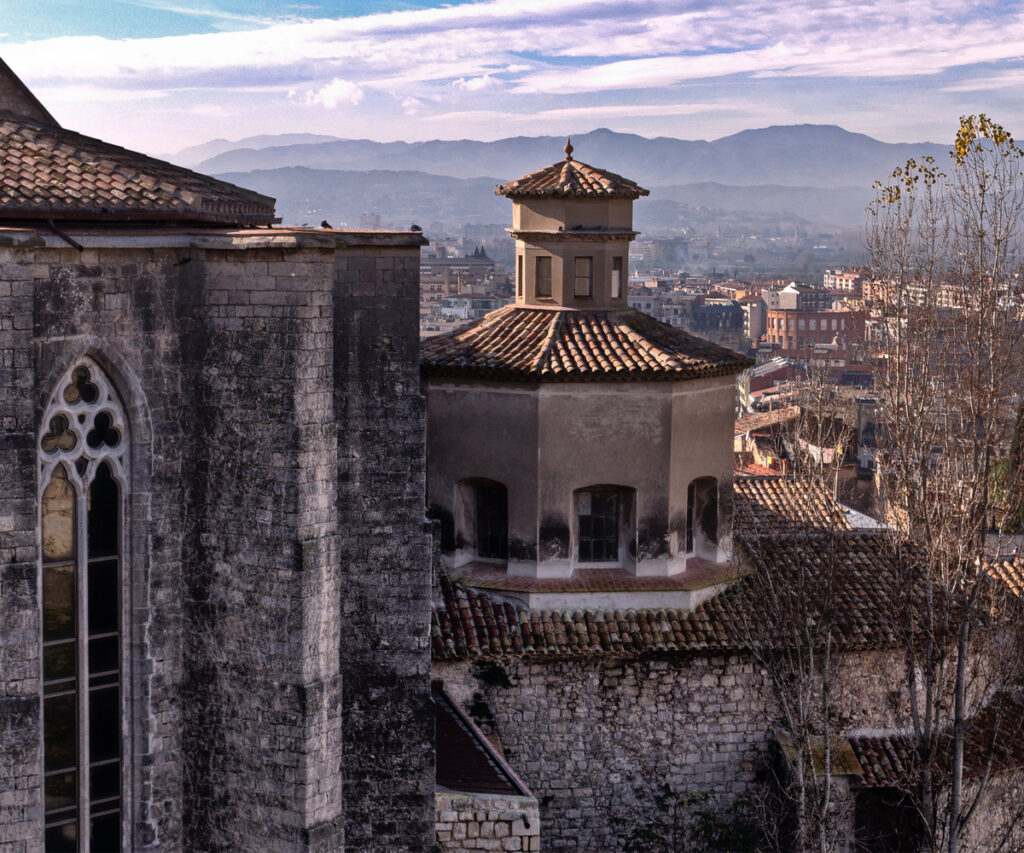 A imagem captura uma vista panorâmica da cidade de Girona, destacando edifícios históricos com arquitetura medieval. Os principais elementos incluem uma torre de pedra com uma janela em arco e um edifício com telhado de telhas. Ao fundo, a paisagem urbana se estende até as montanhas distantes. A cena é banhada por uma luz suave, sugerindo um horário próximo ao amanhecer ou ao pôr do sol.