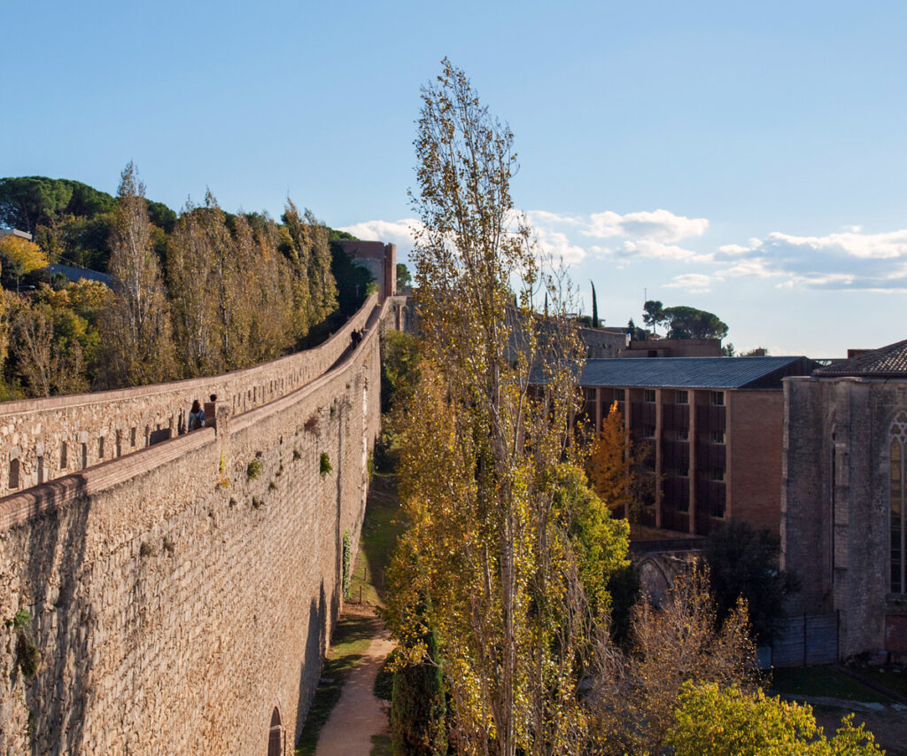 A imagem mostra uma seção da muralha de Girona, uma estrutura histórica que se estende ao longe. Pessoas podem ser vistas caminhando ao longo do topo da muralha. Ao fundo, há árvores com folhas amarelas e laranja, indicando a estação de outono. Edifícios históricos e modernos são visíveis, com um céu azul claro e algumas nuvens adicionando profundidade à cena.