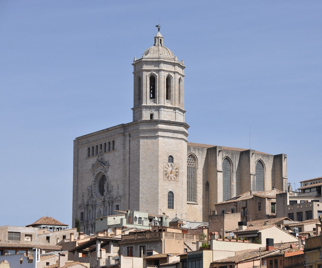 A imagem mostra a Catedral de Girona, um marco histórico com uma torre de relógio proeminente e edifícios antigos ao redor. A catedral é construída com pedra e apresenta um estilo arquitetônico gótico. O céu está limpo e azul, proporcionando um contraste acentuado com a estrutura de pedra.