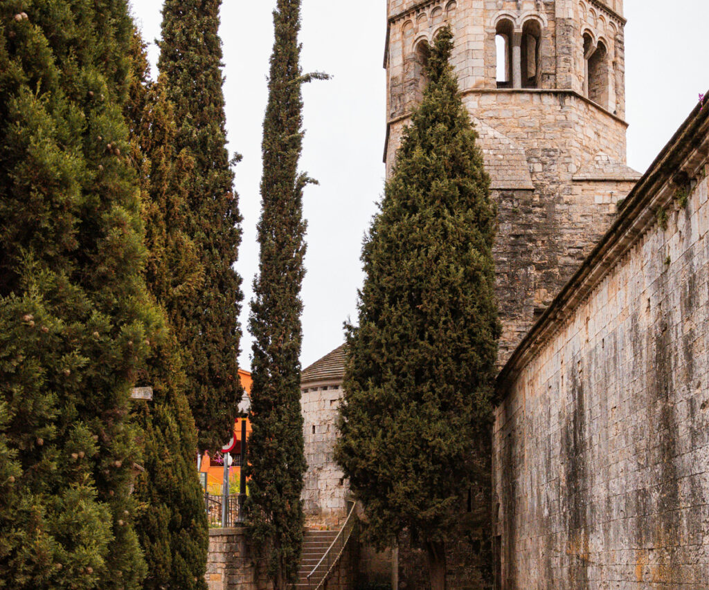 A imagem retrata o Monastério Sant Pere De Galligants, um marco histórico com uma torre de pedra proeminente. A cena é emoldurada por árvores altas e bem cuidadas, criando uma atmosfera serena e majestosa. O monastério é cercado por um muro de pedra, e ao fundo, há um edifício com telhado vermelho e uma escada que leva a ele.