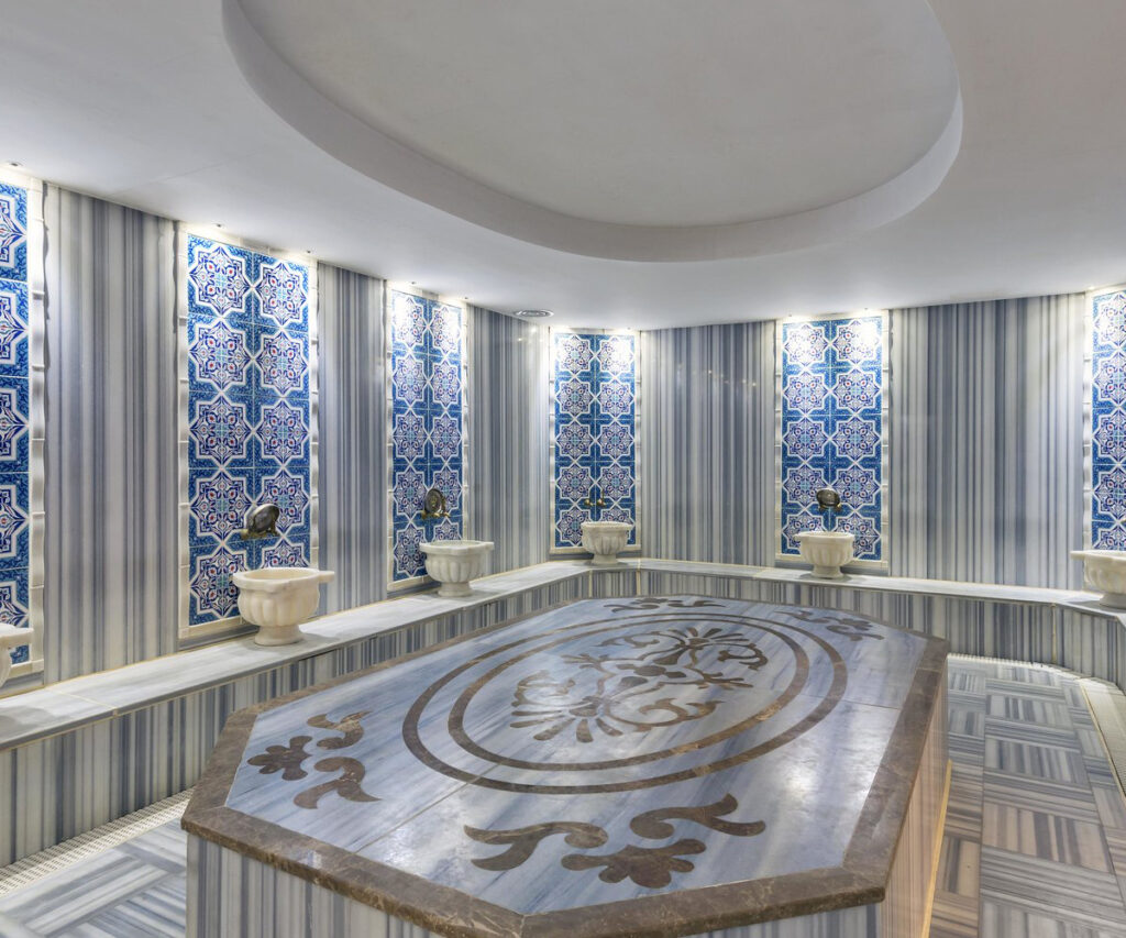 A imagem mostra um spa luxuoso com uma maca de pedra para a prática do Hammam. O ambiente é decorado com azulejos azuis e brancos intricados e cortinas verticais. A maca de pedra no centro é adornada com entalhes dourados e é cercada por vasos de cerâmica branca.
