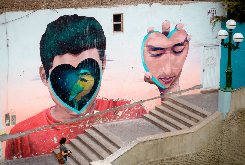 A imagem apresenta um mural de grafite vibrante criado pelo artista Jade Rivera no bairro de Barranco, em Lima, Peru. O mural retrata dois rostos humanos com corações no lugar dos olhos, cada coração abrigando um pássaro colorido. O fundo é uma mistura de tons pastéis, com um rosa suave e um azul claro. O mural está situado em uma parede de um edifício com uma porta branca e um poste de luz próximo. Uma pessoa é vista caminhando na frente do mural, adicionando um senso de escala à obra de arte.